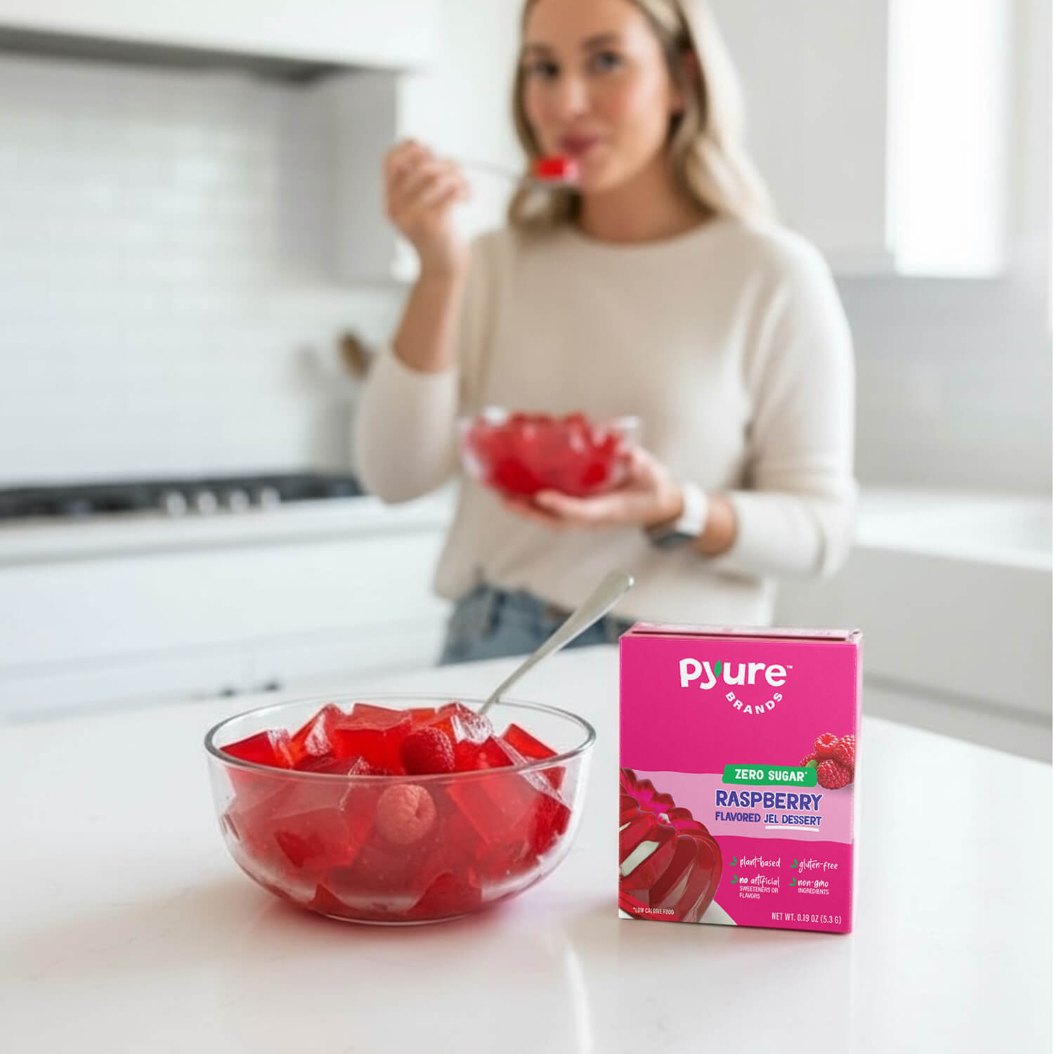 Woman eating raspberry-flavored jel dessert with a box of Pyure jel dessert mix on a kitchen counter.